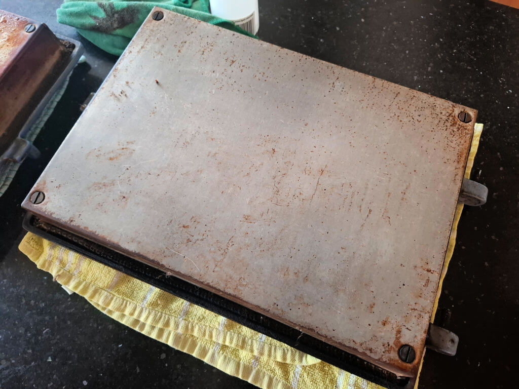 Aga door resting on a yellow tea towel. Its surface is marked with burnt on grease, and there's a green cloth nearby on a dark countertop.