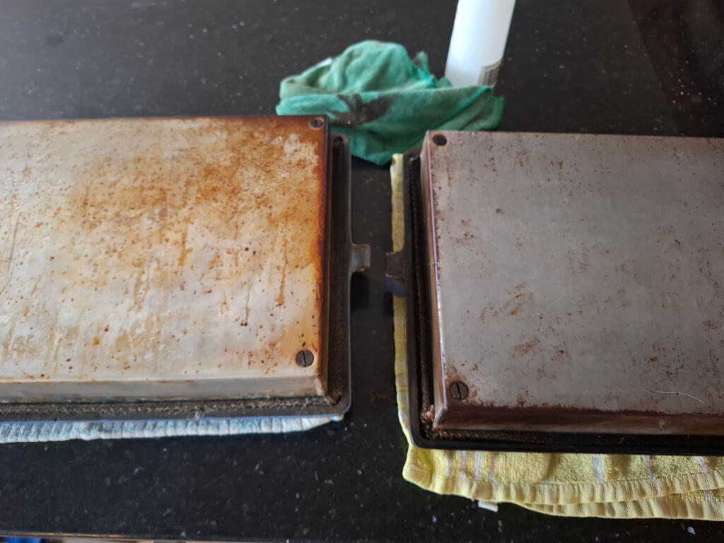 Two metal Aga doors on a black countertop, one with burnt on grease, the other cleaner. Green and yellow cloths in the background convey a cleaning theme.