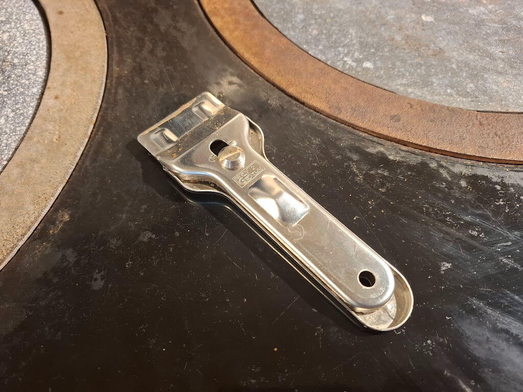 A silver metal scraper lies on a black Aga stovetop, next to hot plates. The scene conveys an industrial, utilitarian mood.