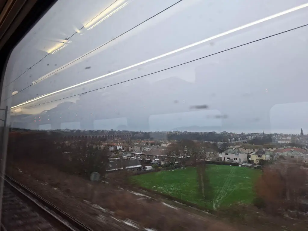 View from a train window on a cloudy day showing a town with a green field. In the distance, an arched viaduct is visible against the grey sky.