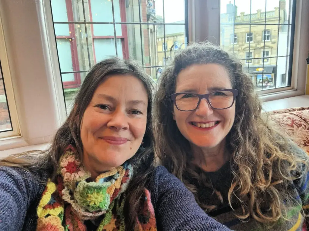 Two women smiling indoors by a window. One wears a colourful scarf, the other glasses. Outside, a cityscape with historic buildings is visible. Warm, cheerful atmosphere.
