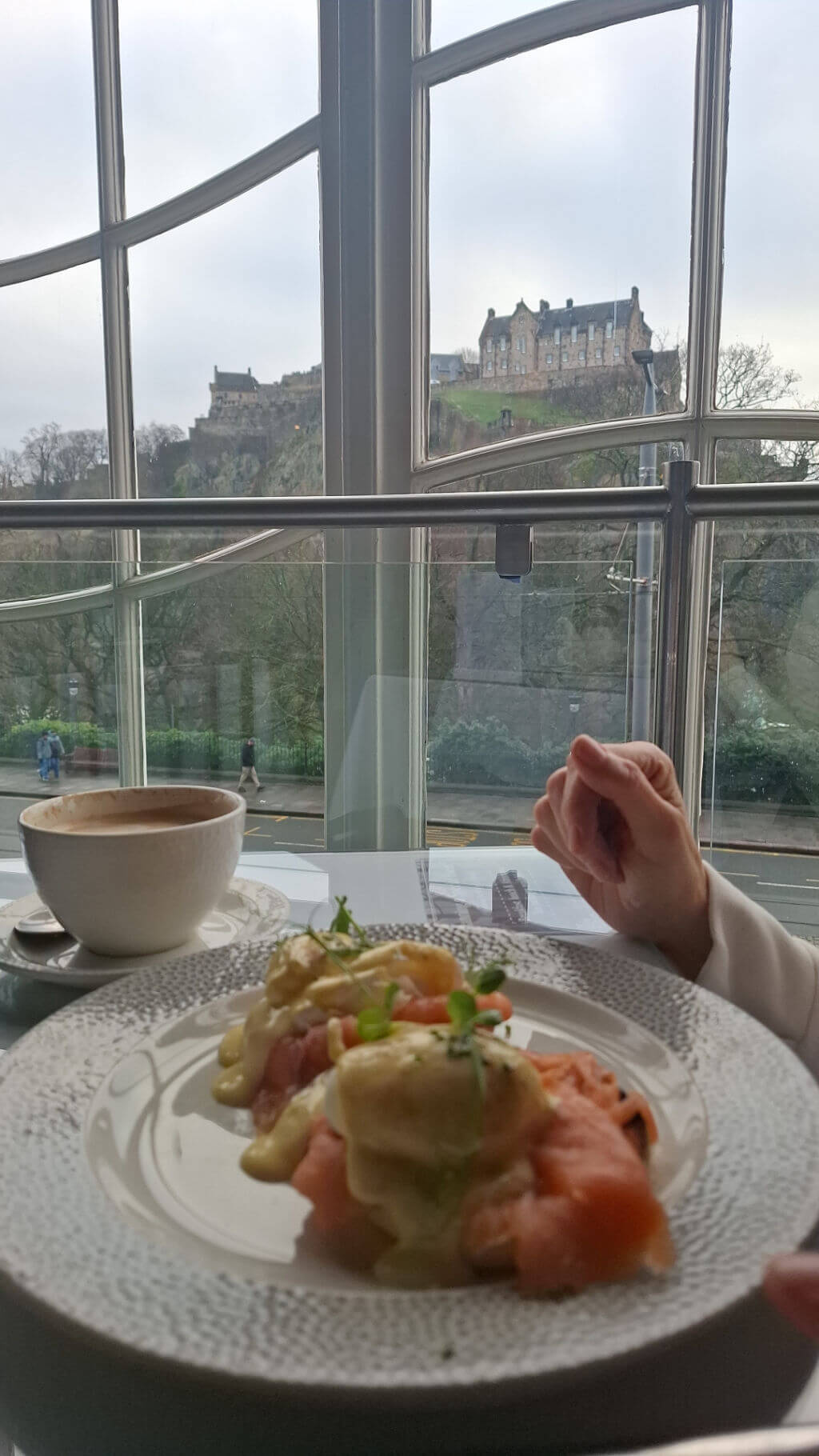 A plate of eggs Royale sits on a table by a large window, with a cappuccino and a distant view of a castle outside.