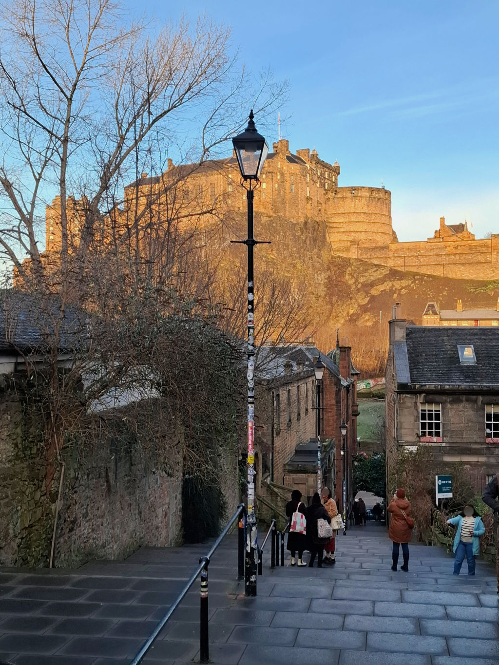 View of Edinburgh Castle from a cobblestone street, with a lamppost in front and people casually walking. The scene is under a clear blue sky.