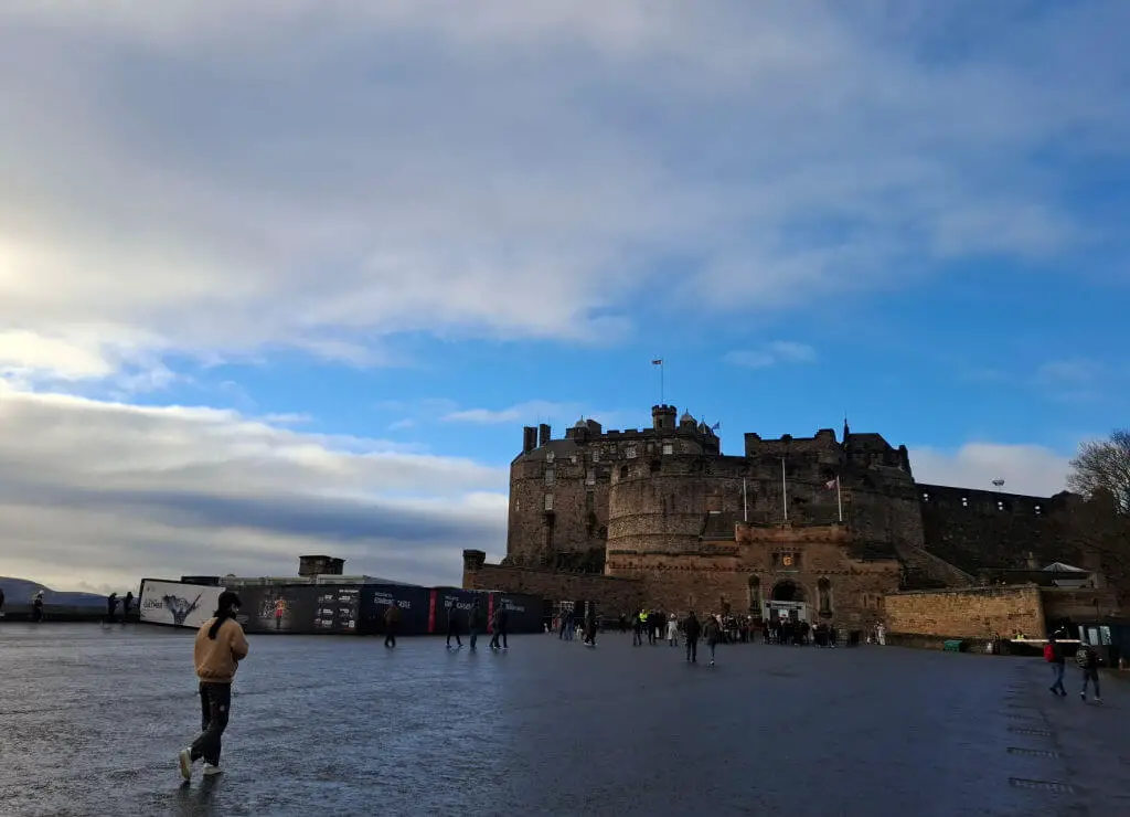 Historic stone castle on a hill under a partly cloudy blue sky. Visitors walk on the wide, open courtyard, creating a lively and inviting scene.