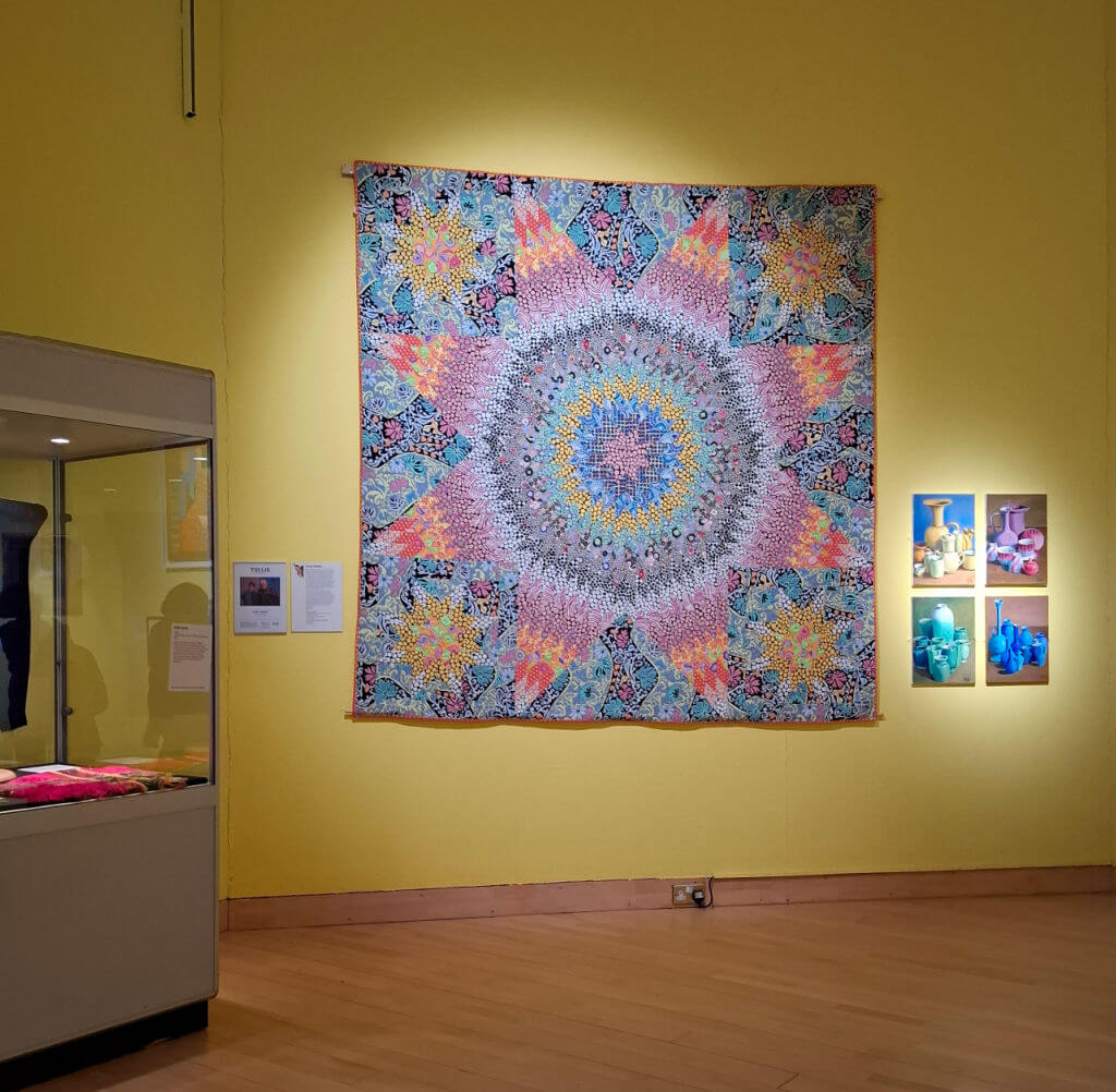 A vibrant quilt with a mandala pattern in shades of pink, blue, and yellow hangs on a yellow wall in a gallery, alongside small colourful photographs.