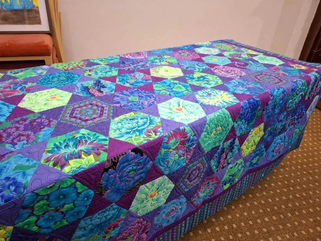 Colourful quilt draped over a table, featuring vibrant geometric patterns in blues, purples, and greens. The intricate design creates a lively, cosy tone.