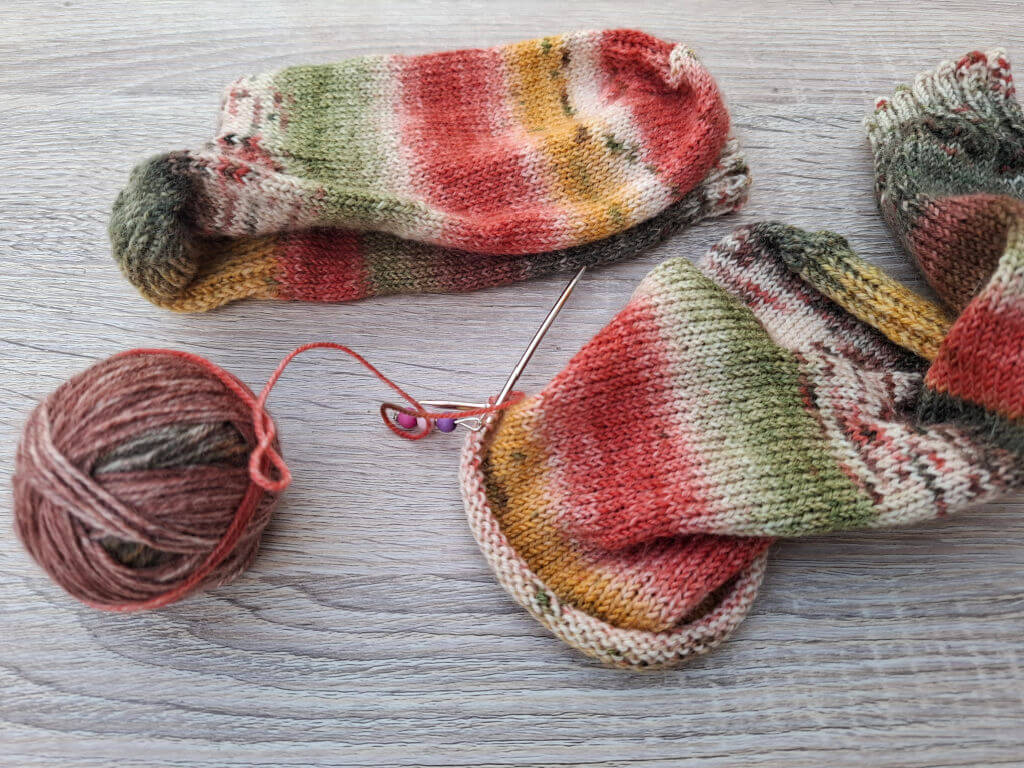 Multicoloured knitted socks with red, green, and orange stripes lie on a wooden surface. A yarn ball and knitting needles complete the cosy scene.