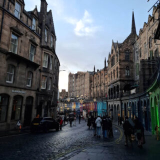 A charming cobblestone street lined with historic buildings and colourful shopfronts. People walk under a clear sky, creating a lively, inviting atmosphere.