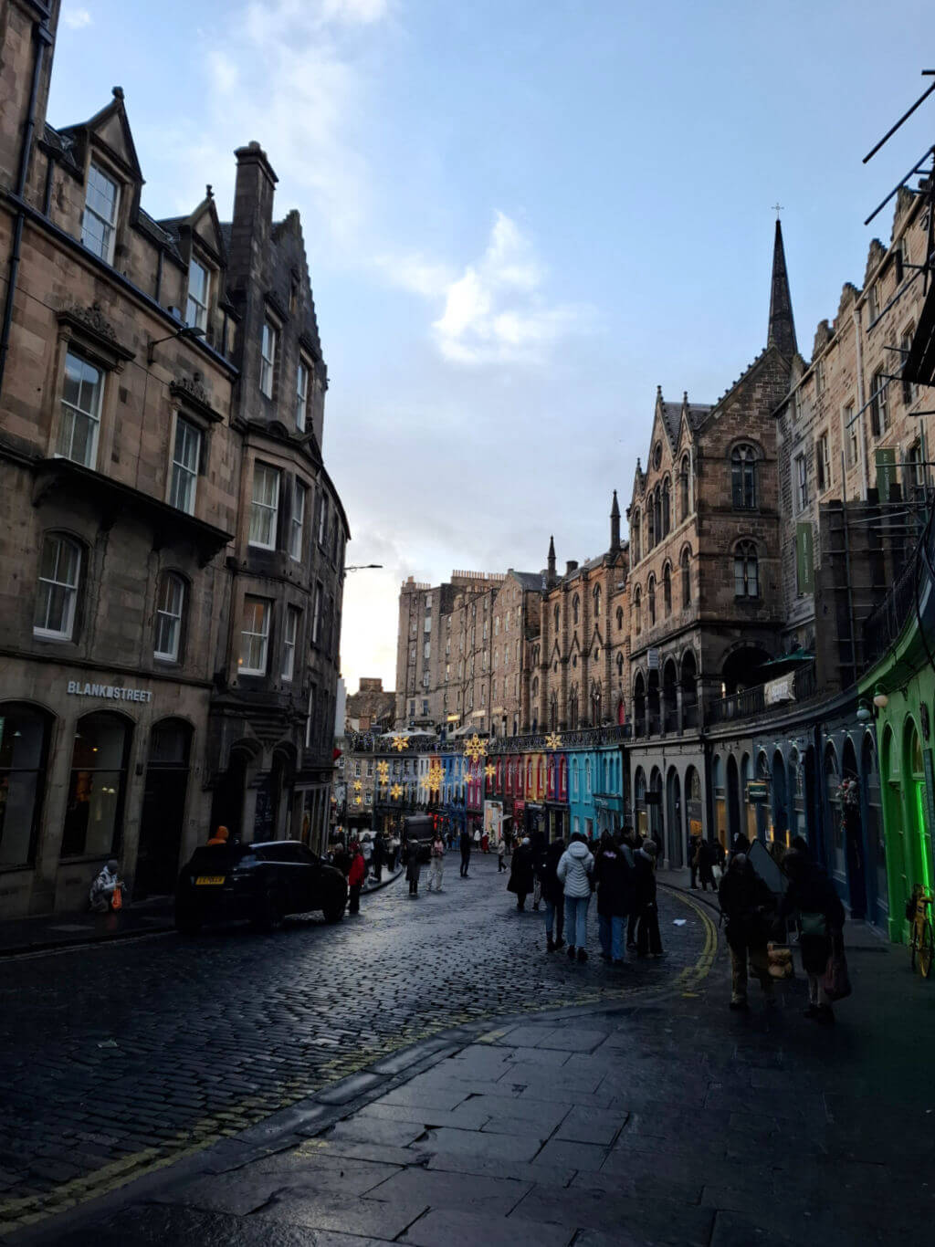 A charming cobblestone street lined with historic buildings and colourful shopfronts. People walk under a clear sky, creating a lively, inviting atmosphere.