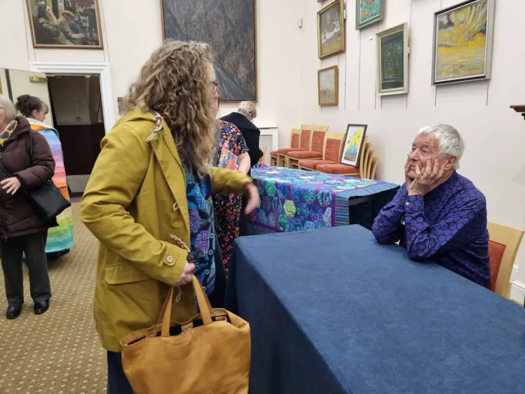 A woman in a yellow coat interacts with an elderly man seated at a table in a gallery. Artworks hang on the walls, creating a warm, contemplative atmosphere.