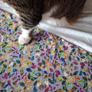 A cat's front legs and white paws are resting on a colourful, leaf-patterned fabric. The playful design contrasts with the cat's striped fur.