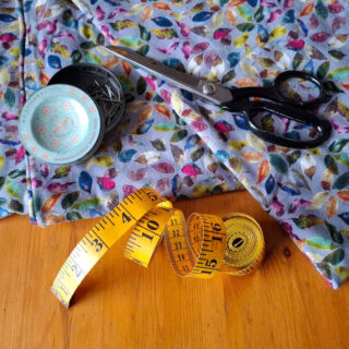 A vibrant, leaf-patterned fabric lies on a wooden table with sewing tools. Black scissors, a yellow measuring tape, and a tin of pins convey a crafting theme.