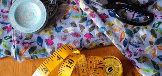 A vibrant, leaf-patterned fabric lies on a wooden table with sewing tools. Black scissors, a yellow measuring tape, and a tin of pins convey a crafting theme.