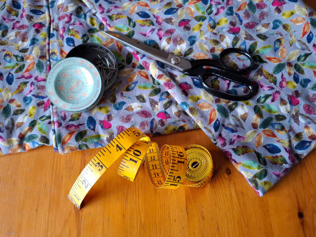 A vibrant, leaf-patterned fabric lies on a wooden table with sewing tools. Black scissors, a yellow measuring tape, and a tin of pins convey a crafting theme.