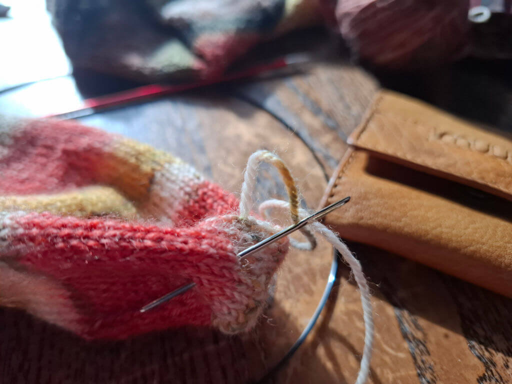 Close-up of a knitting project with a threaded needle in colourful yarn. A leather pouch and blurred knitting in the background. Warm, cosy atmosphere.