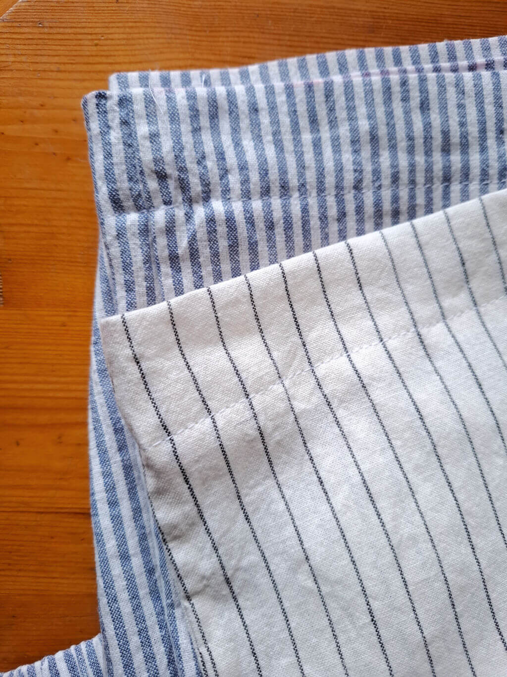 Two pairs of trousers are on a wooden surface showing the hemline stitching. One is blue with white stripes, and the other is white with thin black stripes, creating a cosy, rustic feel.