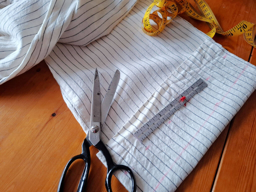 Scissors and a metal ruler lie on a striped trouser leg on a wooden table. A yellow measuring tape is curled nearby, suggesting sewing preparations.