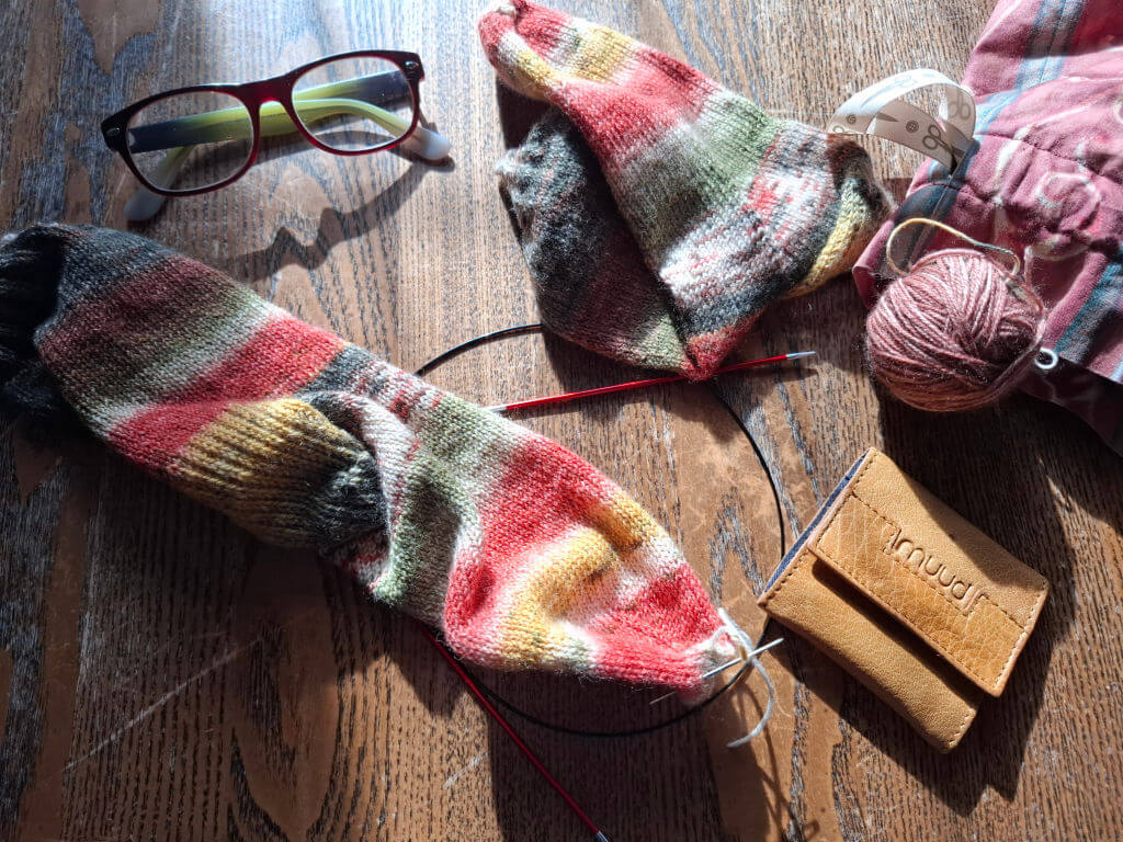 A knitting project with multicoloured yarn, partially completed socks on needles, glasses, yarn ball, project bag, and a leather case on a wooden table.