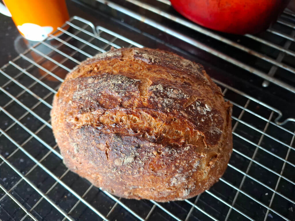 A round, crusty loaf of brown bread cools on a wire rack, with a golden-brown, cracked surface, conveying warmth and freshness in a cosy kitchen.