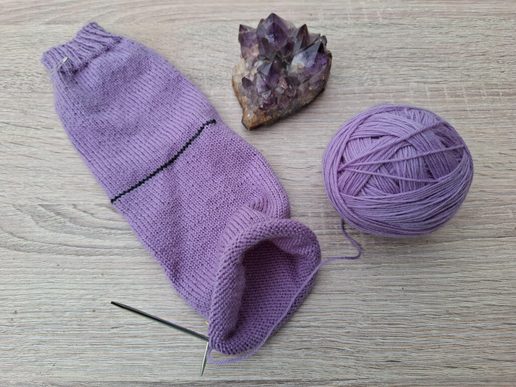 A purple knit sock with a black stripe is partially completed, lying on a wooden surface. Nearby are a ball of matching yarn and an amethyst crystal.