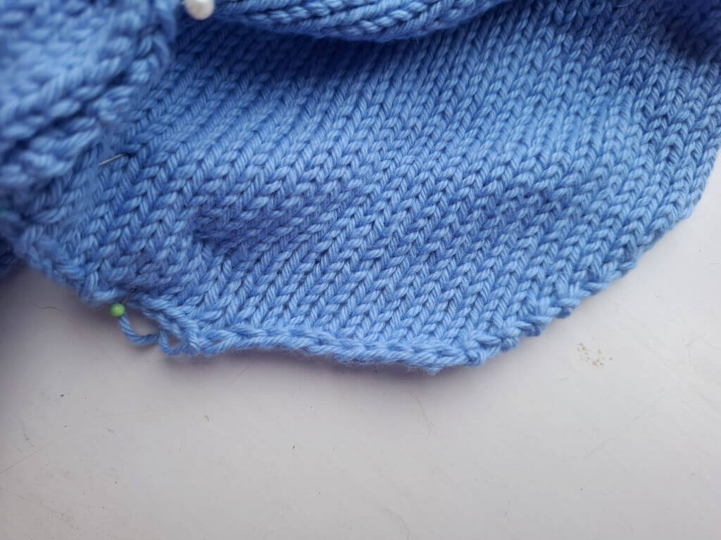 Close-up of a light blue knitted fabric with a smooth stockinette stitch pattern. The texture is soft, and the edges are slightly curled.