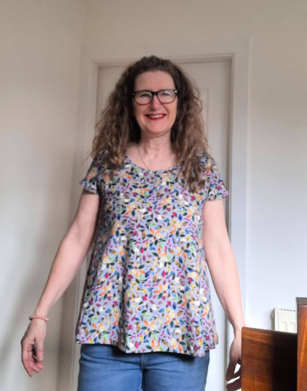 A woman with long curly hair and glasses smiles warmly. She wears a colourful floral shirt and jeans, standing in a bright room with a wooden table.