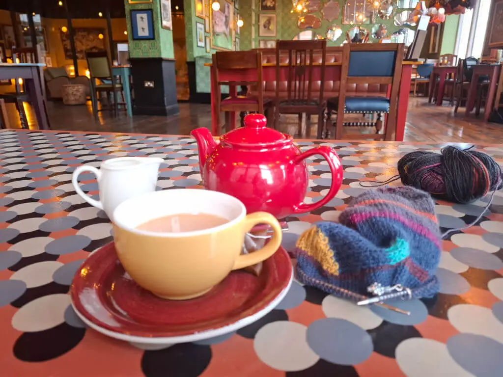 A cosy café scene with a red teapot, a yellow cup of tea, cream, and knitting yarn on a polka-dotted table. Warm lighting and wooden chairs in the background.