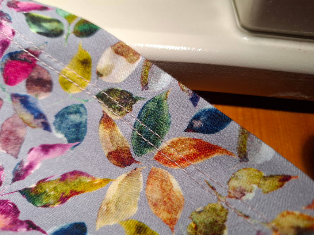 Close-up of a colorful fabric with a leaf pattern, featuring vibrant hues like green, blue, and pink on a light background. Twin stitching is visible, creating a neat, orderly design, conveying a sense of craft and creativity.