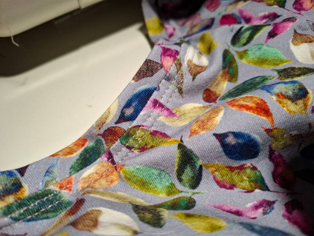 Close-up of twin stitches along the neckline of a colourful fabric with a leaf pattern in vibrant orange, green, blue, and pink hues on a light grey background, evoking a cheerful tone.
