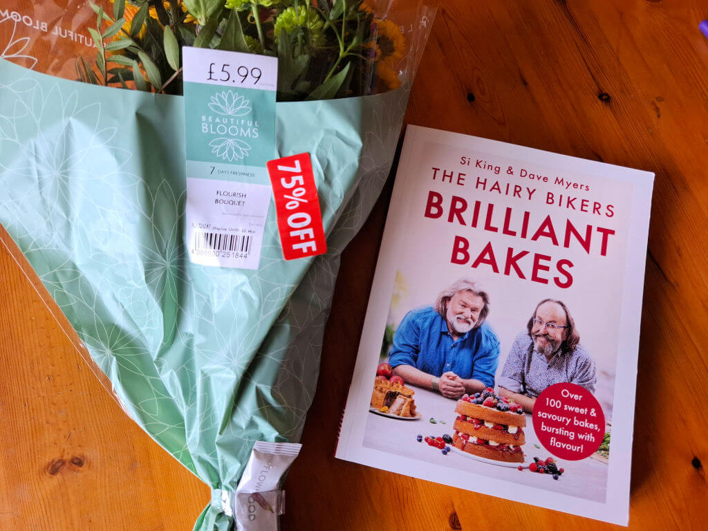 A bouquet wrapped in mint paper with a 75% off tag beside a cookbook titled "The Hairy Bikers Brilliant Bakes" on a wooden table.