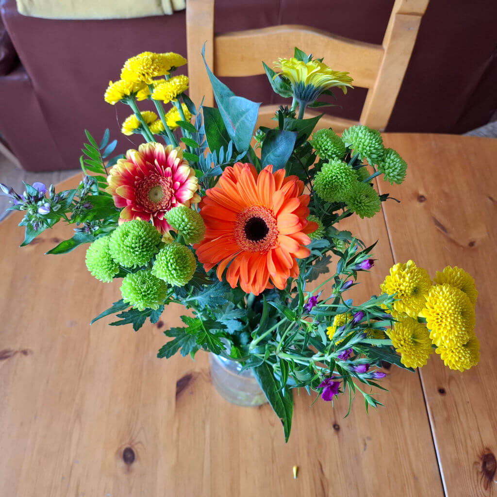 A colourful flower arrangement in a vase on a wooden table. Features orange gerbera, yellow and green chrysanthemums, and multicoloured blooms. Bright and cheerful ambiance.