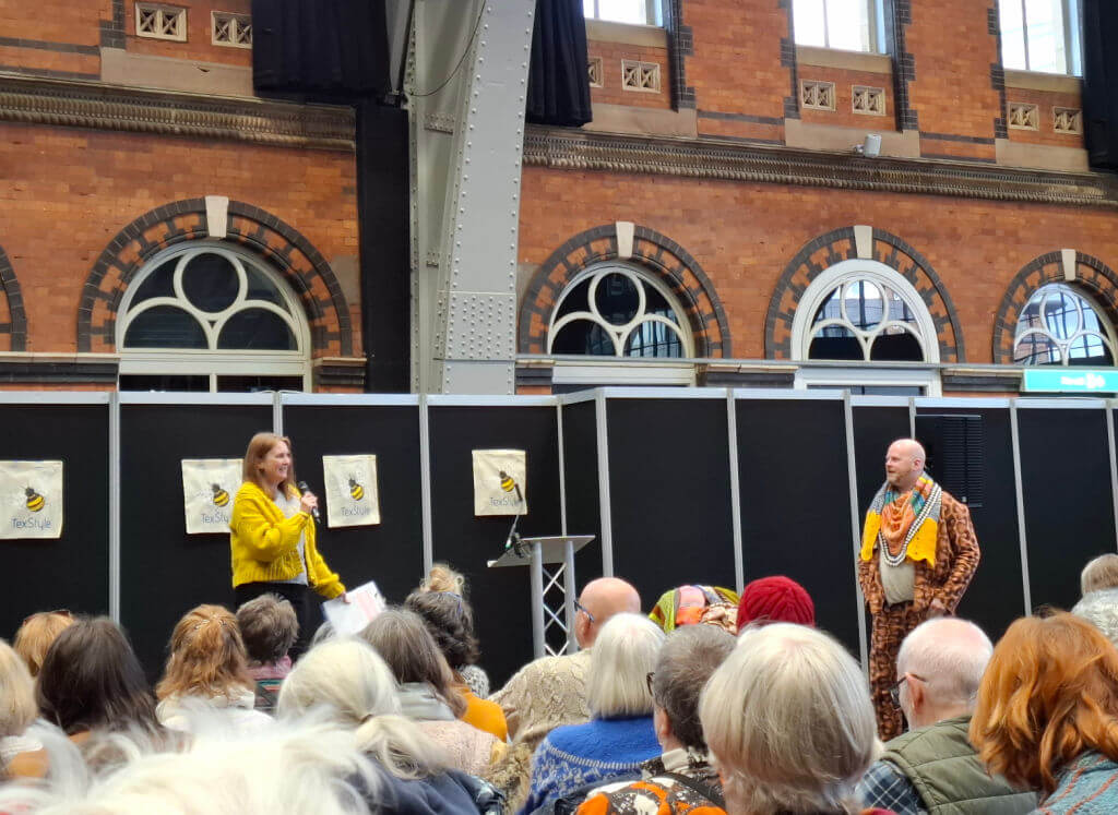 A woman in a yellow sweater speaks into a microphone on a stage beside a man in patterned attire. They face an audience seated in a historical venue with arched windows and brick walls. The scene is lively and engaging.