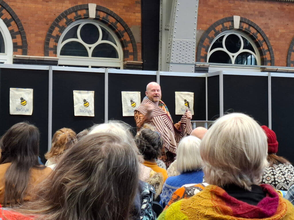 A man in a striped shawl speaks to an attentive audience inside a brick-walled venue with arched windows, creating a warm, engaging atmosphere.