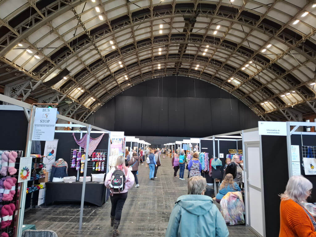 A large convention hall filled with people browsing craft stalls. The arched metal ceiling and bright lights create an inviting atmosphere, with vibrant colours of yarn and fabric visible at the booths.
