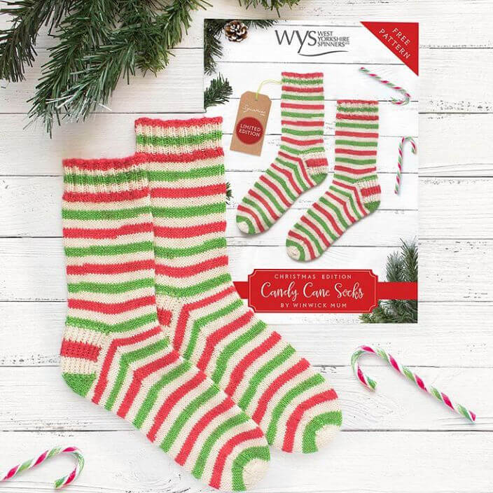 Striped socks in red, green, and white lie on a white wooden surface, mimicking a candy cane pattern. Beside them, a holiday-themed packaging. Festive vibe.