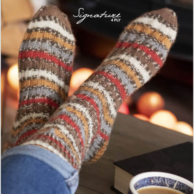 Cosy knitted socks with a striped pattern in warm tones rest on a table near a lit fireplace. A book and a cup of tea add to the relaxing atmosphere.