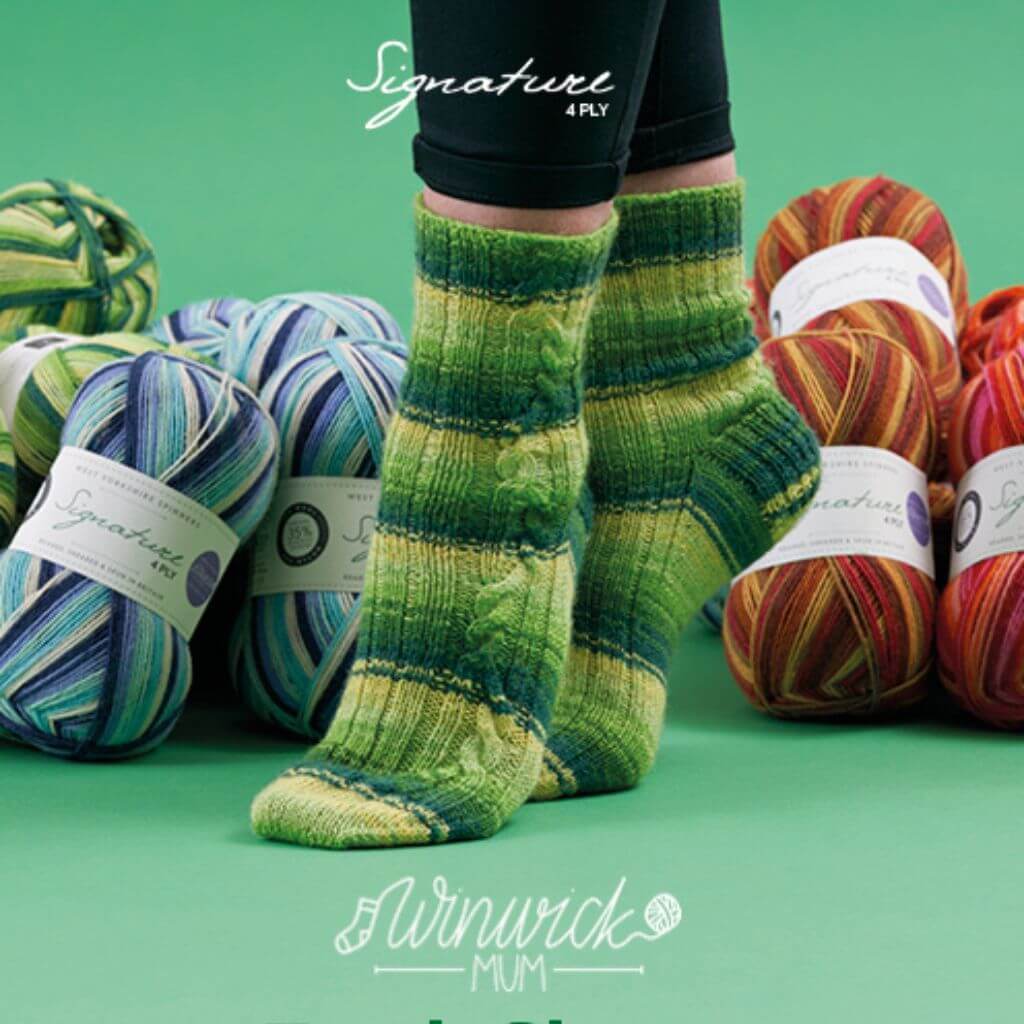 Person wearing green striped knitted socks stands on a green surface, surrounded by colourful yarn skeins. Brands "Signature" and "Winwick Mum" are visible.