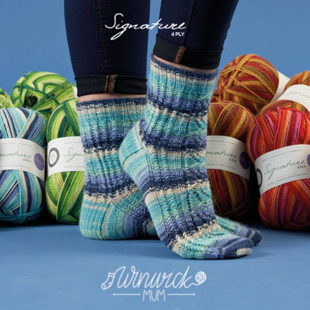 A pair of feet wearing blue striped knitted socks stand on a blue surface. Surrounding them are colourful balls of yarn in green, blue, and red tones. The atmosphere is cosy and creative. Text above reads "Signature 4 Ply," and below is the logo "Winwick Mum."