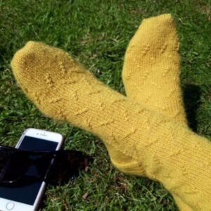 A pair of hand knitted socks in yellow yarn with a twisted stitch pattern. The model is sitting on the grass with feet crossed, and there is a mobile phone and pair of sunglasses next to the feet.
