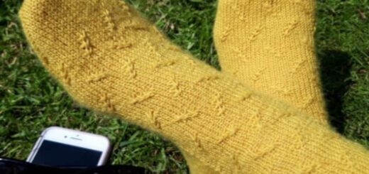 A pair of hand knitted socks in yellow yarn with a twisted stitch pattern. The model is sitting on the grass with feet crossed, and there is a mobile phone and pair of sunglasses next to the feet.