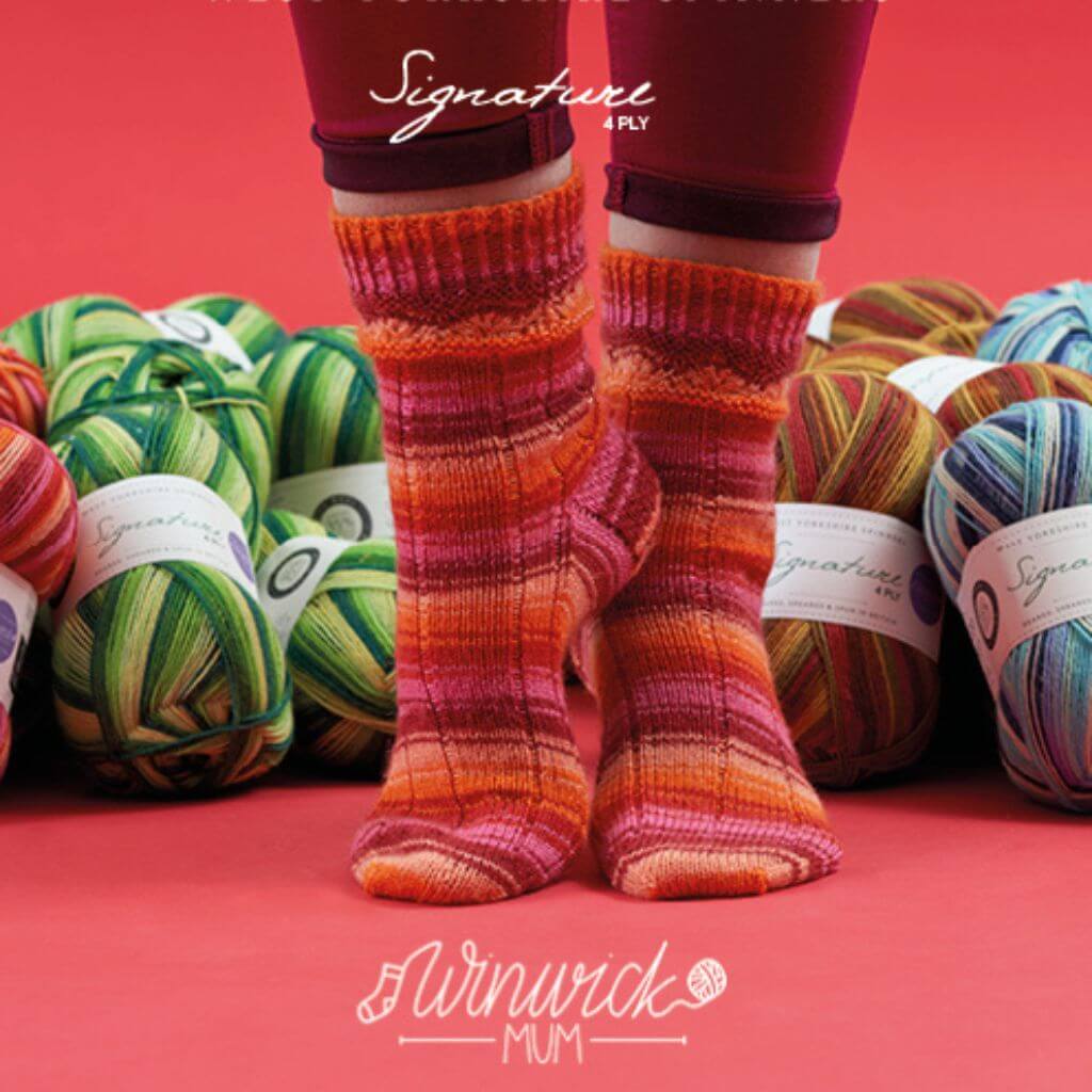 Person wearing cosy, striped pink knit socks stands on a red floor, surrounded by vibrant balls of yarn in green, red, and blue hues. The scene is warm and inviting.