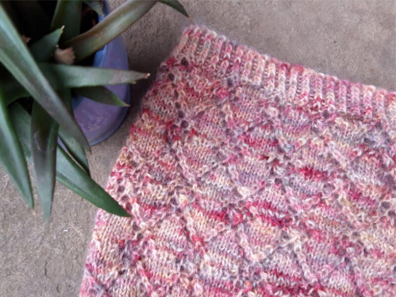 Close-up of a pink and red knitted fabric with a textured diamond pattern, next to a green potted plant in a blue pot on a concrete surface.