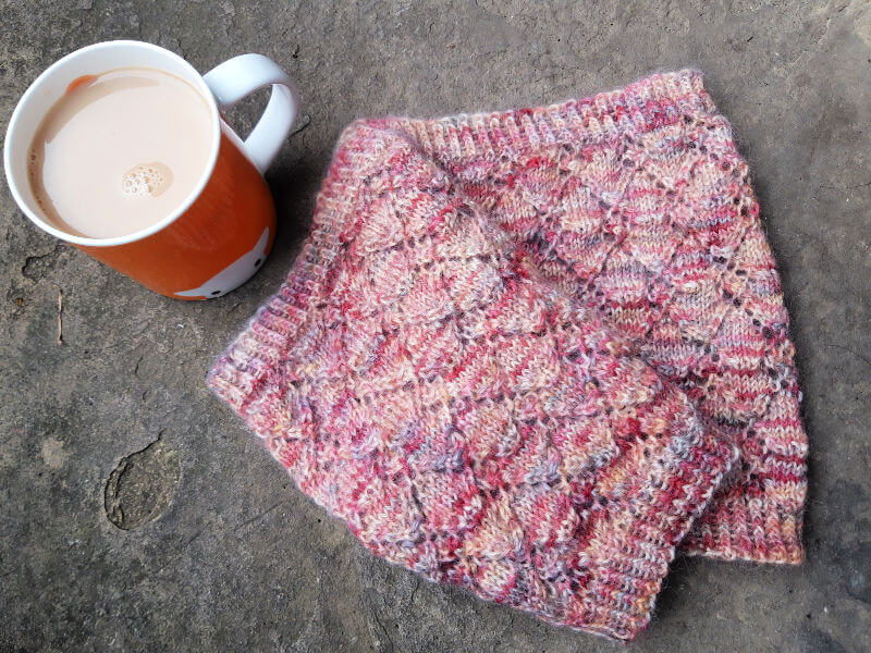 A variegated pink knitted cowl with a diamond pattern lies next to an orange mug filled with tea. The scene is cosy and inviting.
