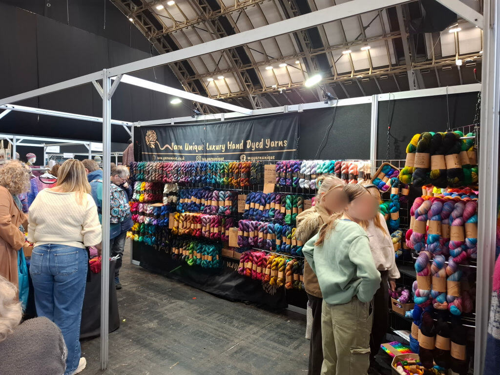 A bustling yarn booth at a craft fair displays vibrant, hand-dyed skeins in various colours. People browse the selection with interest and curiosity.