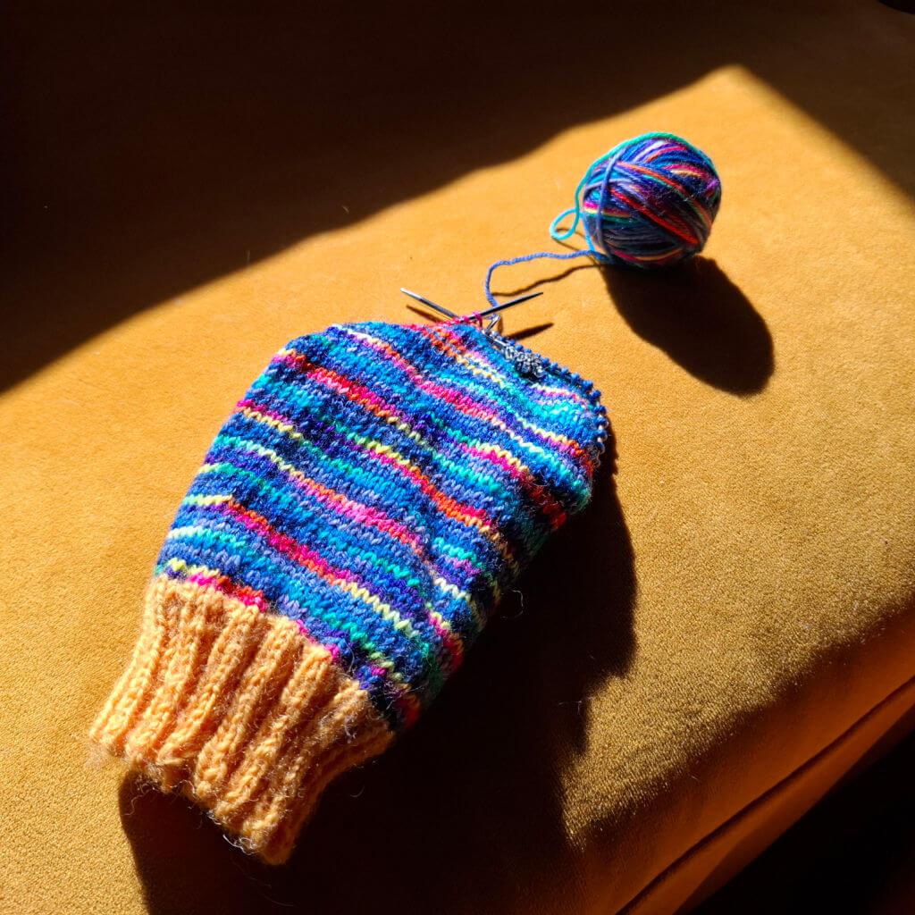 A partly-knitted multicoloured sock with knitting needles, lying on a sunlit sofa. A ball of yarn, casting a shadow, is next to the sock.