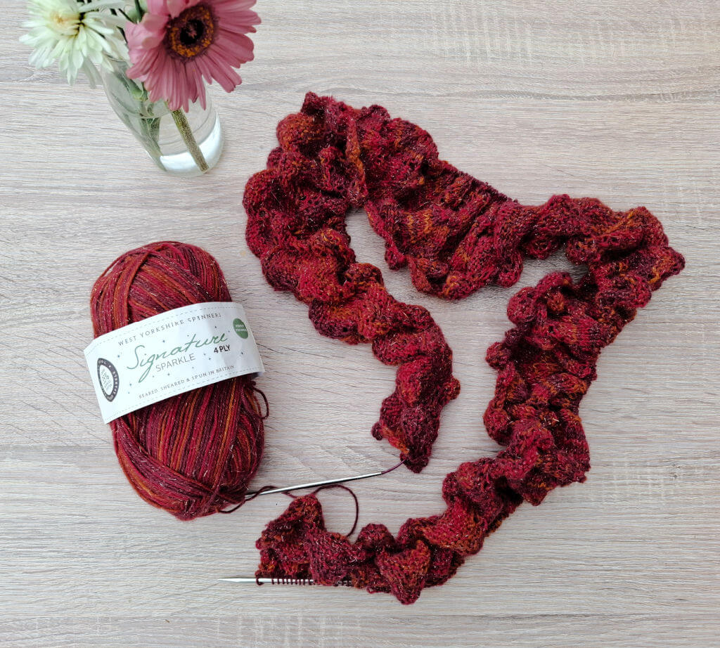A vibrant red sparkly knitted shawl in progress lies on a wooden surface, beside a matching yarn ball. A vase holds pink and white flowers.