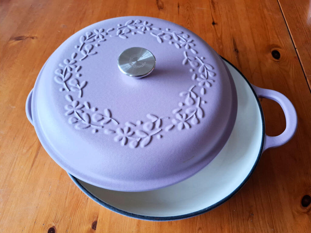 A lavender Dutch oven with a floral pattern on the lid and a shiny silver knob sits on a wooden table. The pot's interior is white.