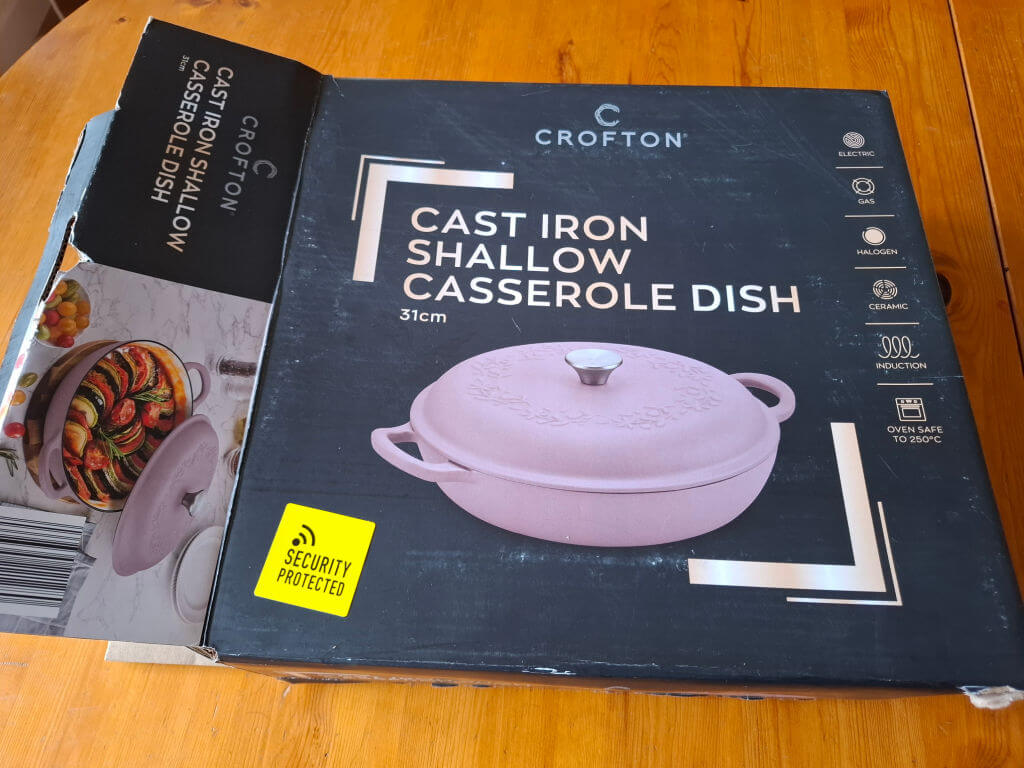 A boxed 31cm Crofton cast iron shallow casserole dish in pink. The box features an image of the dish and icons for electric, gas, halogen, and induction compatibility. It is oven safe up to 250°C. The words 'Security Protected' are on a yellow label. The tone is practical and informative.