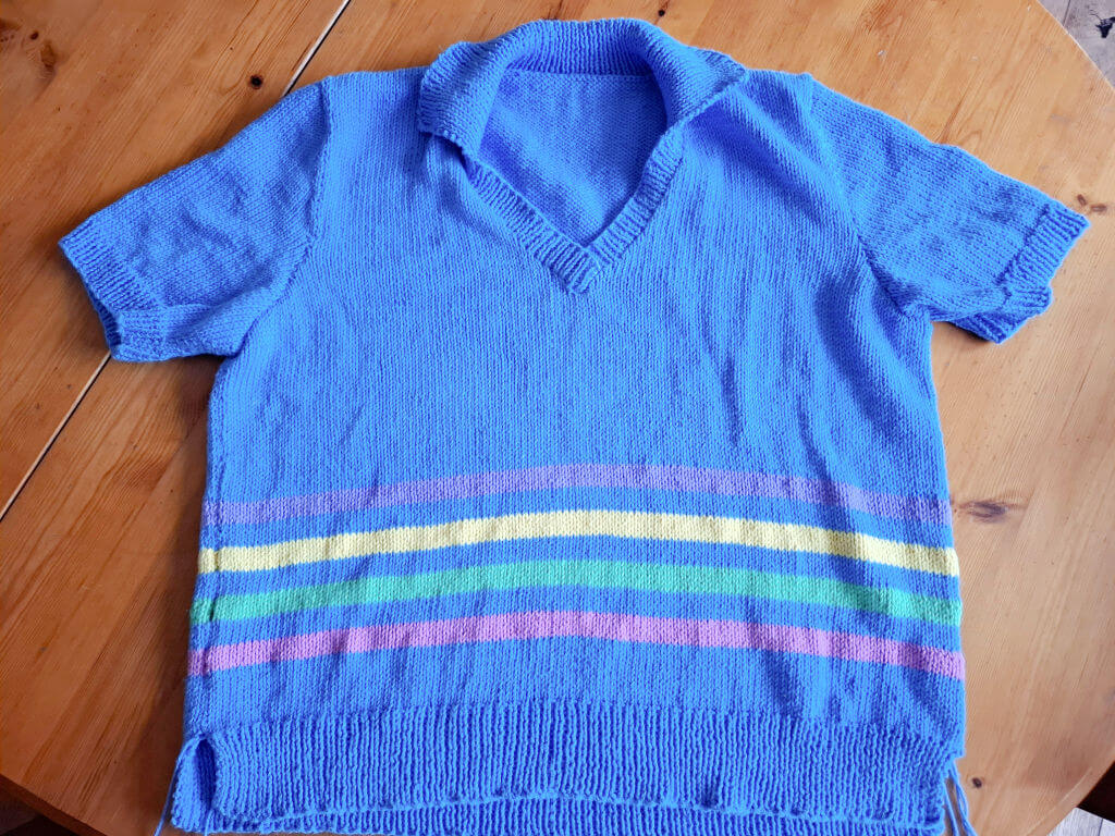 Blue knitted short-sleeve sweater with V-neck, featuring horizontal stripes in pink, yellow, green, and purple, laid flat on a wooden surface.