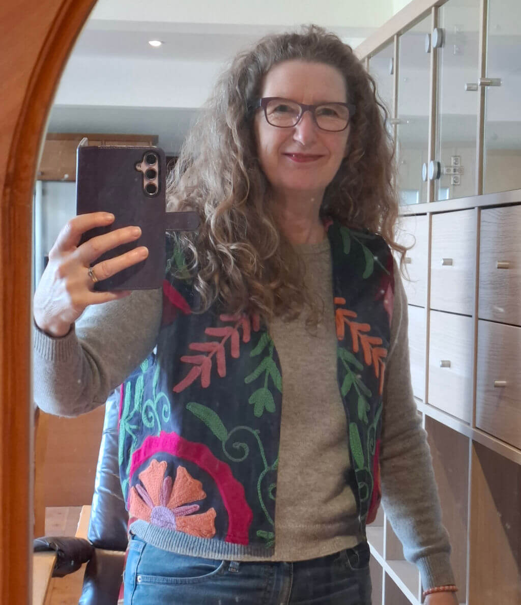 A woman with curly hair takes a mirror selfie, smiling. She wears glasses, a gray sweater, and a colourful embroidered waistcoat. Background: a room with shelves.
